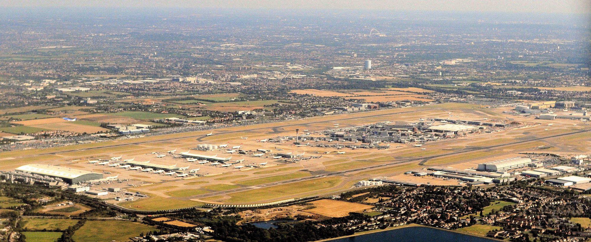 Aerial view of Heathrow Airport showing the terminals, both runways, and aircraft at the stands, with London beyond.