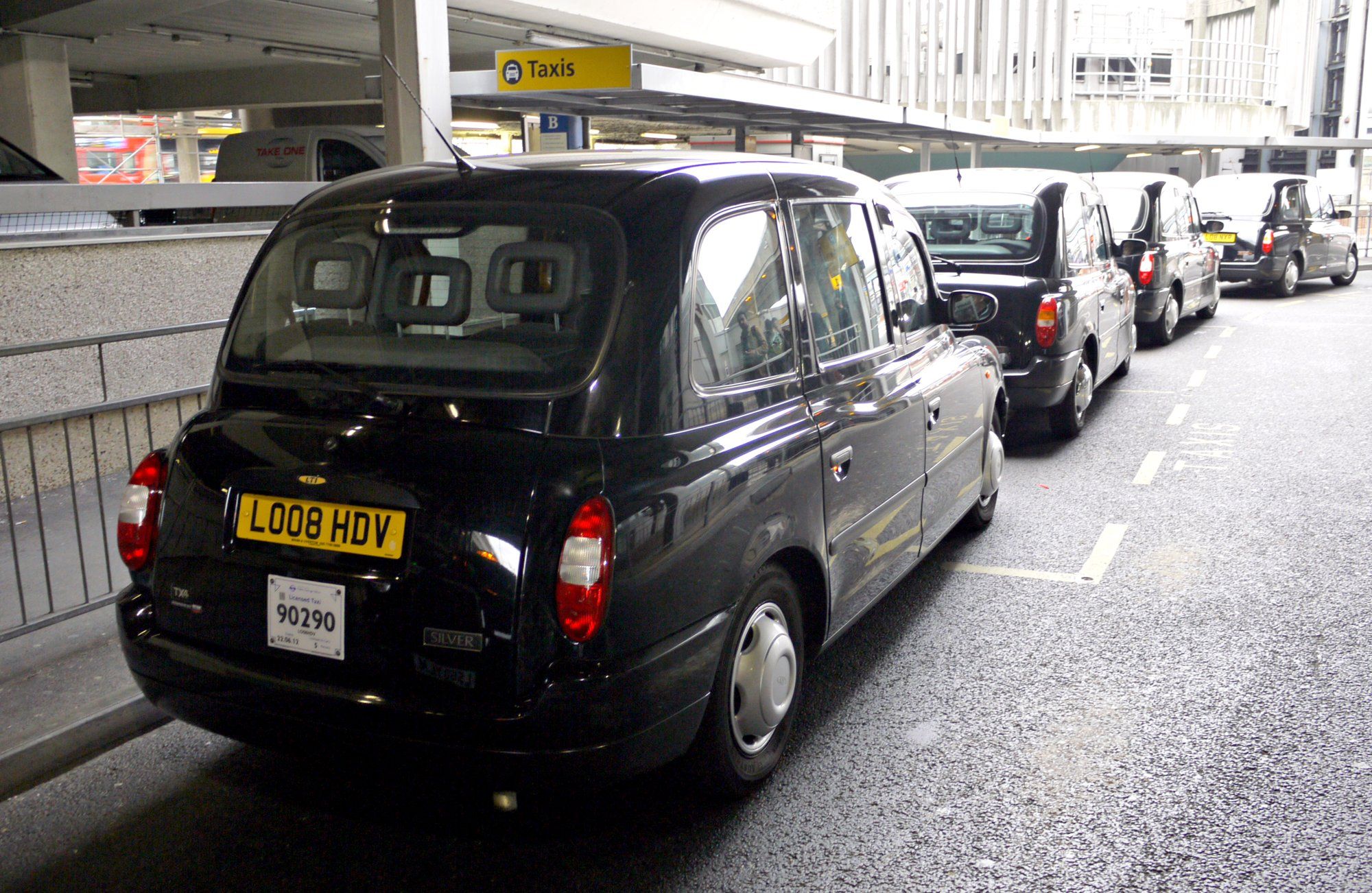 A line of black cabs waiting at the licensed taxi rank outside a Heathrow terminal, under the Taxis sign.