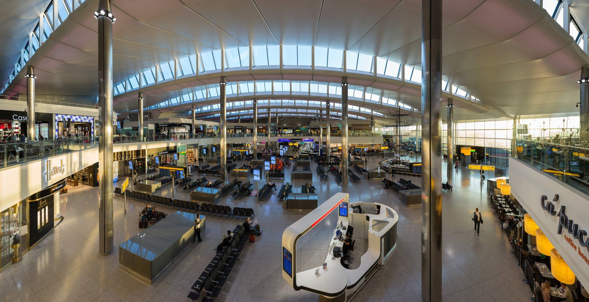 The departures concourse inside Heathrow Terminal 2, the Queen's Terminal, with shops beneath a curved roof.