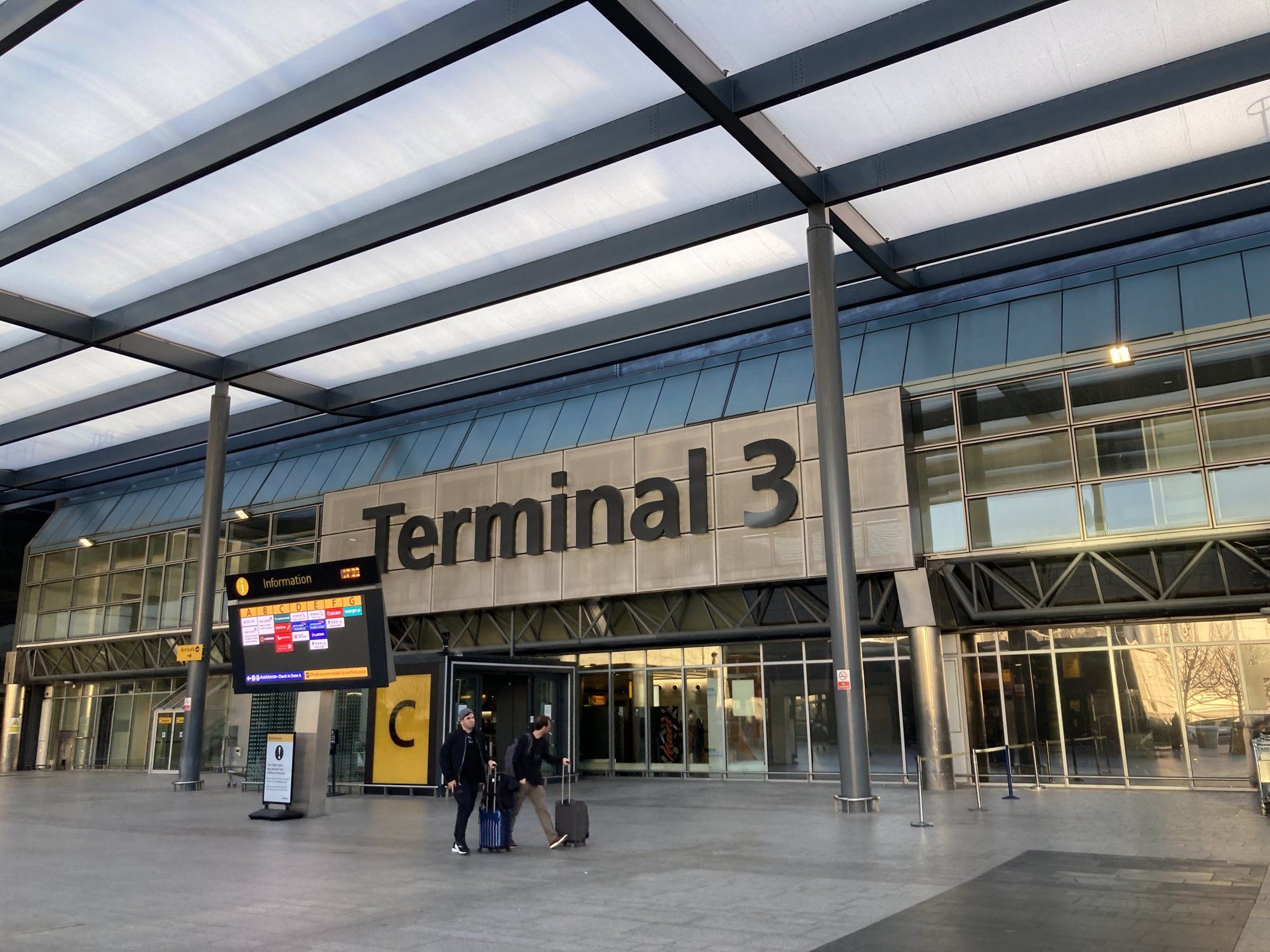 The forecourt entrance to Heathrow Terminal 3, with the Terminal 3 sign above the doors and travellers with luggage.