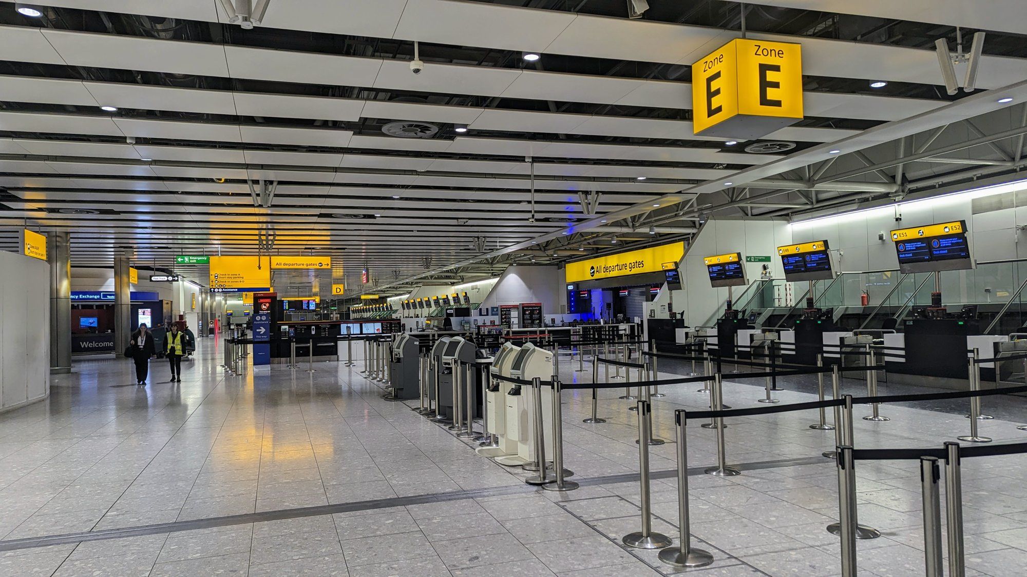 The check-in hall at Heathrow Terminal 4, with self-service kiosks, bag-drop desks, and Zone E signage overhead.
